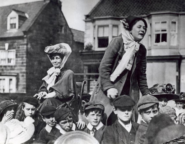Twee suffragettes staan op een kar. Deze vrouwen waren militante voorstandsters van het vrouwenkiesrecht rond 1900. Rondom hen jongens met petjes. Waarschijnlijk in Engeland.Two suffragettes (women's rights movement) standing on a cart, bringing their message. England, location unknown, about 1900.